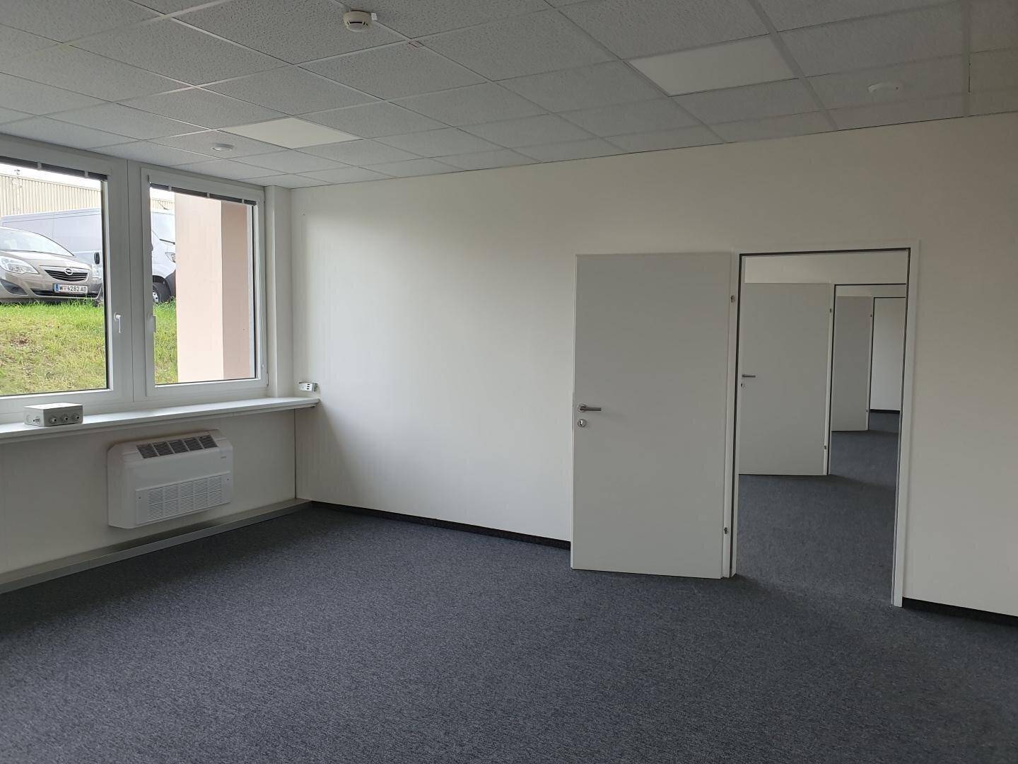 Geräumiges Büro mit Teppichboden, großen Fenstern und Zugang zu weiteren Räumen.