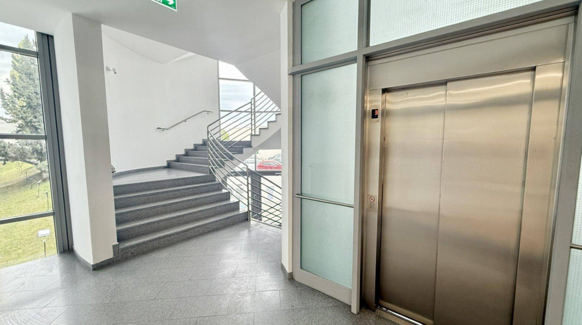 Heller Eingangsbereich mit moderner Treppe, großen Fenstern und einem Aufzug.