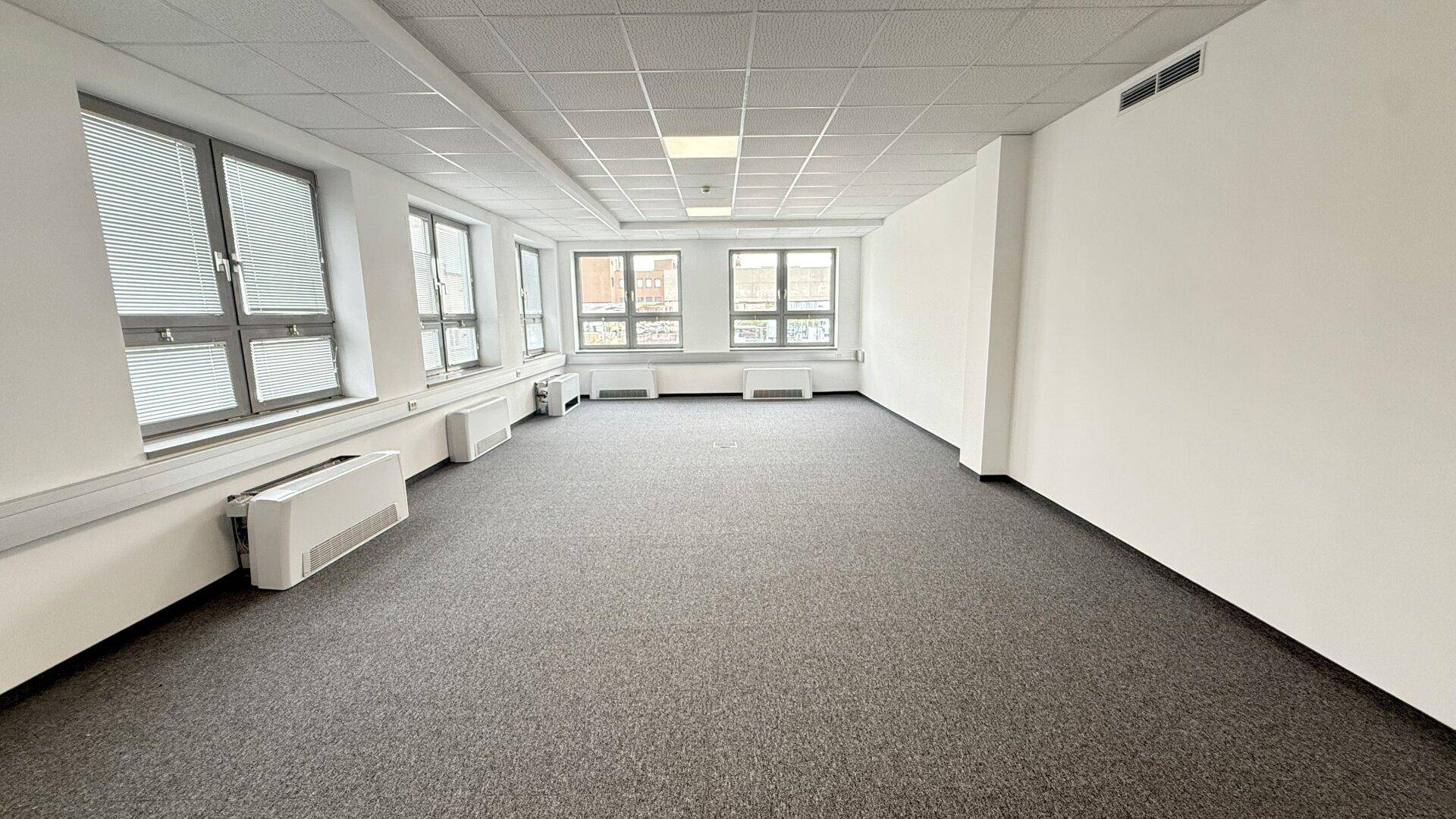 Helles und geräumiges Büro mit Teppichboden, großen Fenstern und Jalousien für angenehmes Arbeiten.