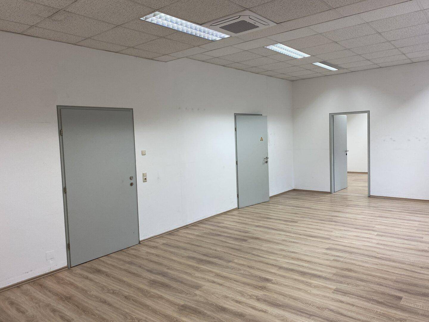 Heller Büroraum mit Laminatboden, weißen Wänden und mehreren Türen zu angrenzenden Bereichen.
