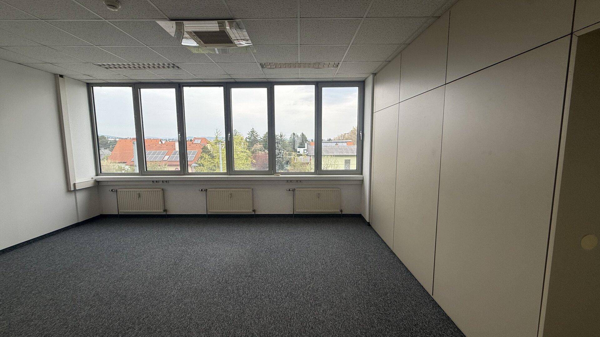 Leeres Büro mit großen Fenstern, Teppichboden und Heizkörpern unter den Fenstern.