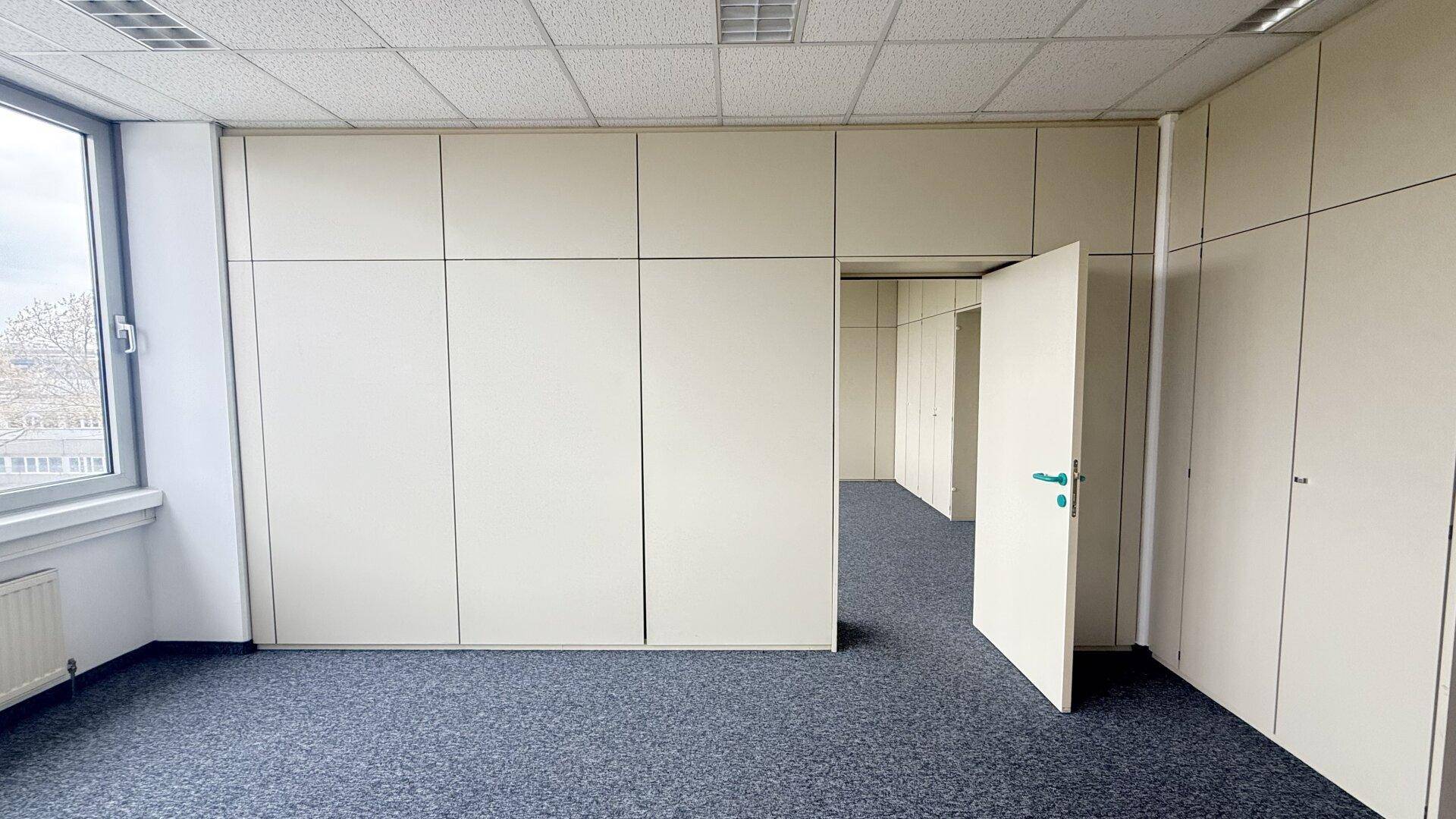 Büroraum mit modularen Trennwänden, Teppichboden und einer offenen Tür zu einem weiteren Bereich.