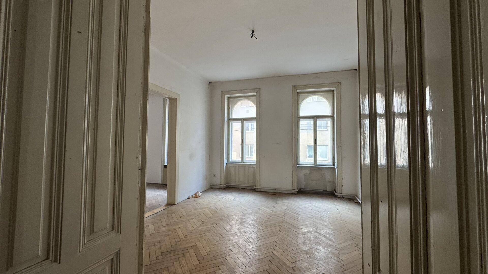 Leeres Wohnzimmer mit Fischgrätparkett und hohen Fenstern, bereit zur Renovierung in einem Altbau.