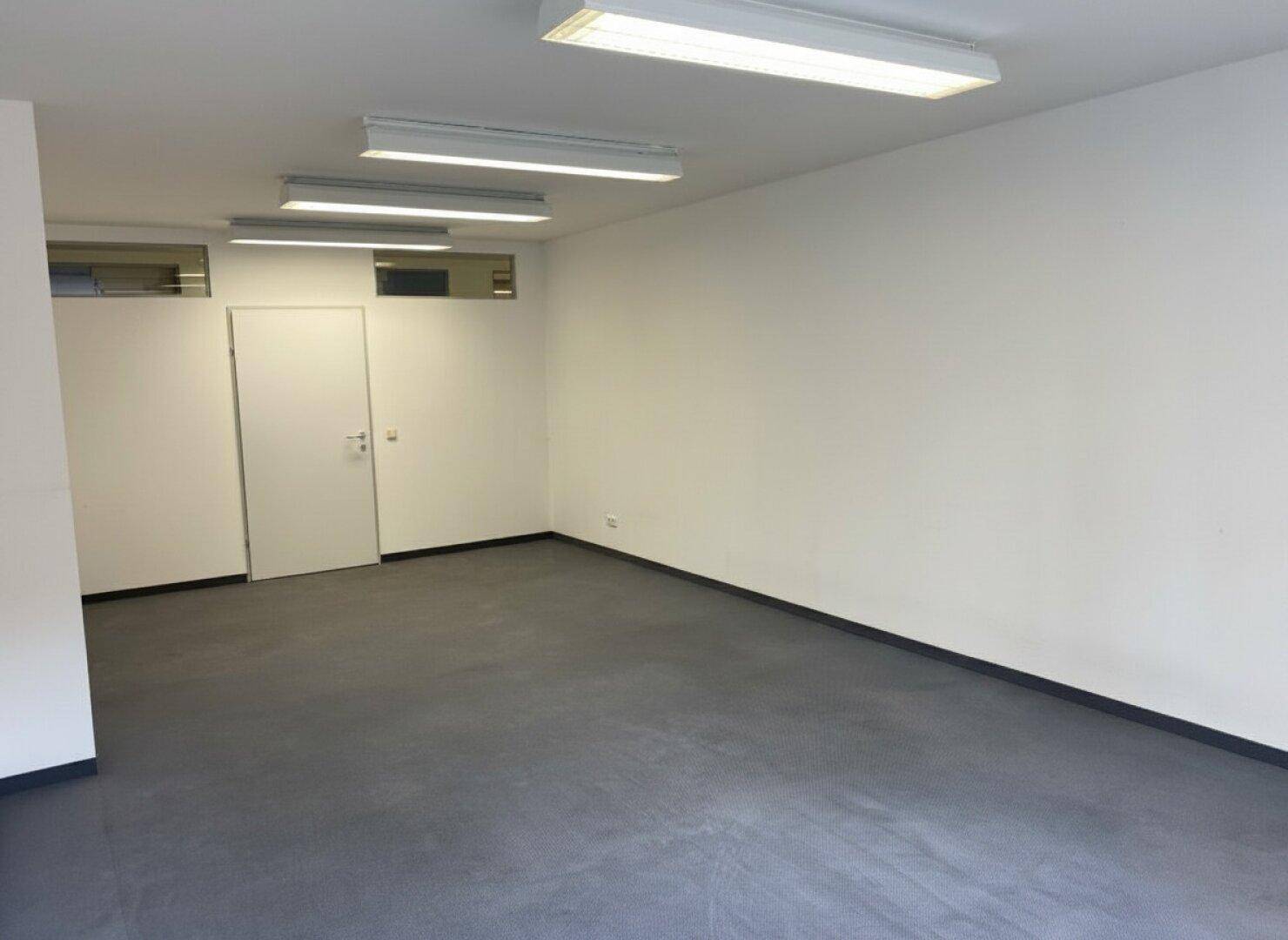 Geräumiger Büroraum mit weiß gestrichenen Wänden, dunklem Vinylboden und Deckenleuchten.