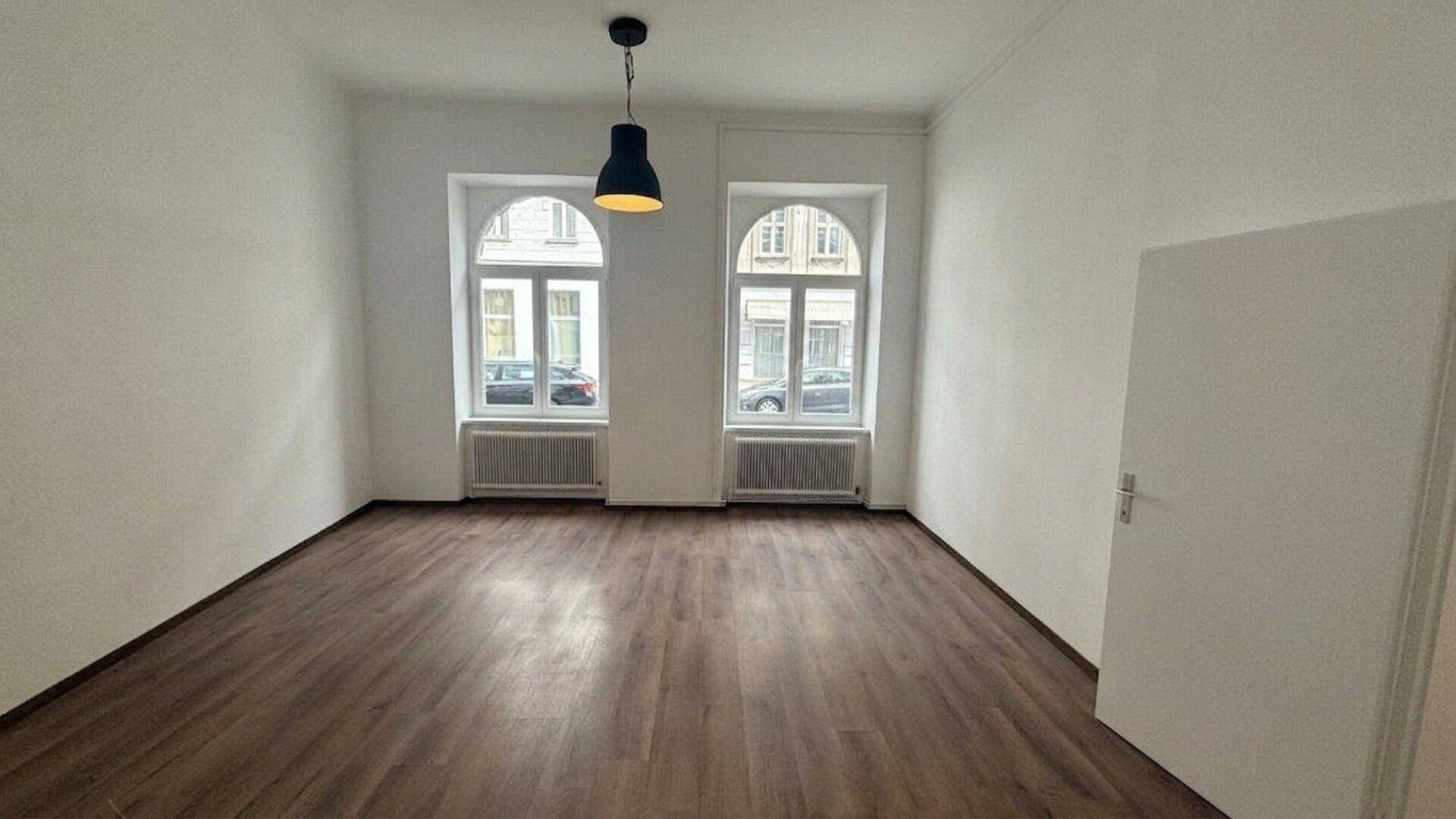 Geräumiger, unmöblierter Raum mit Laminatboden, weißen Wänden und zwei hohen Fenstern.