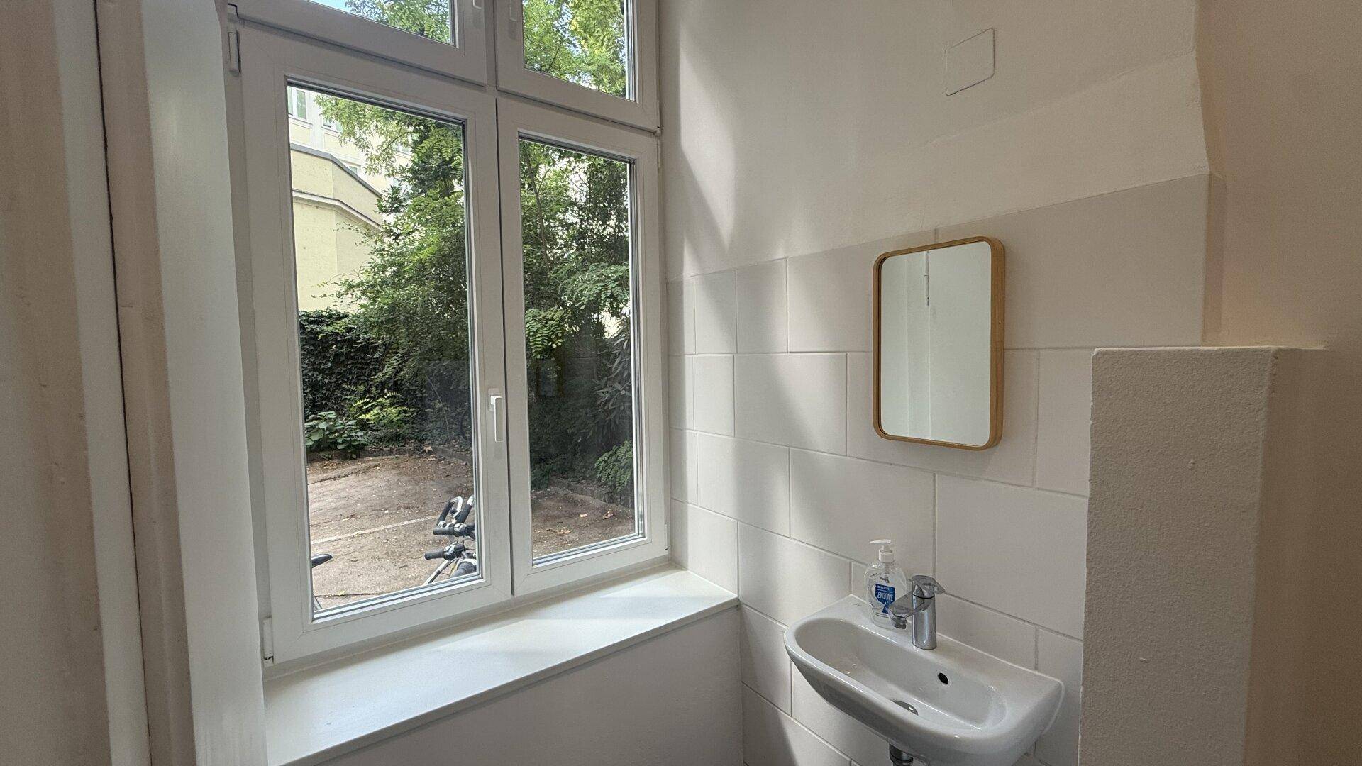 Kleines Badezimmer mit Fenster, Waschbecken und Spiegel, teilweise gefliesten Wänden.