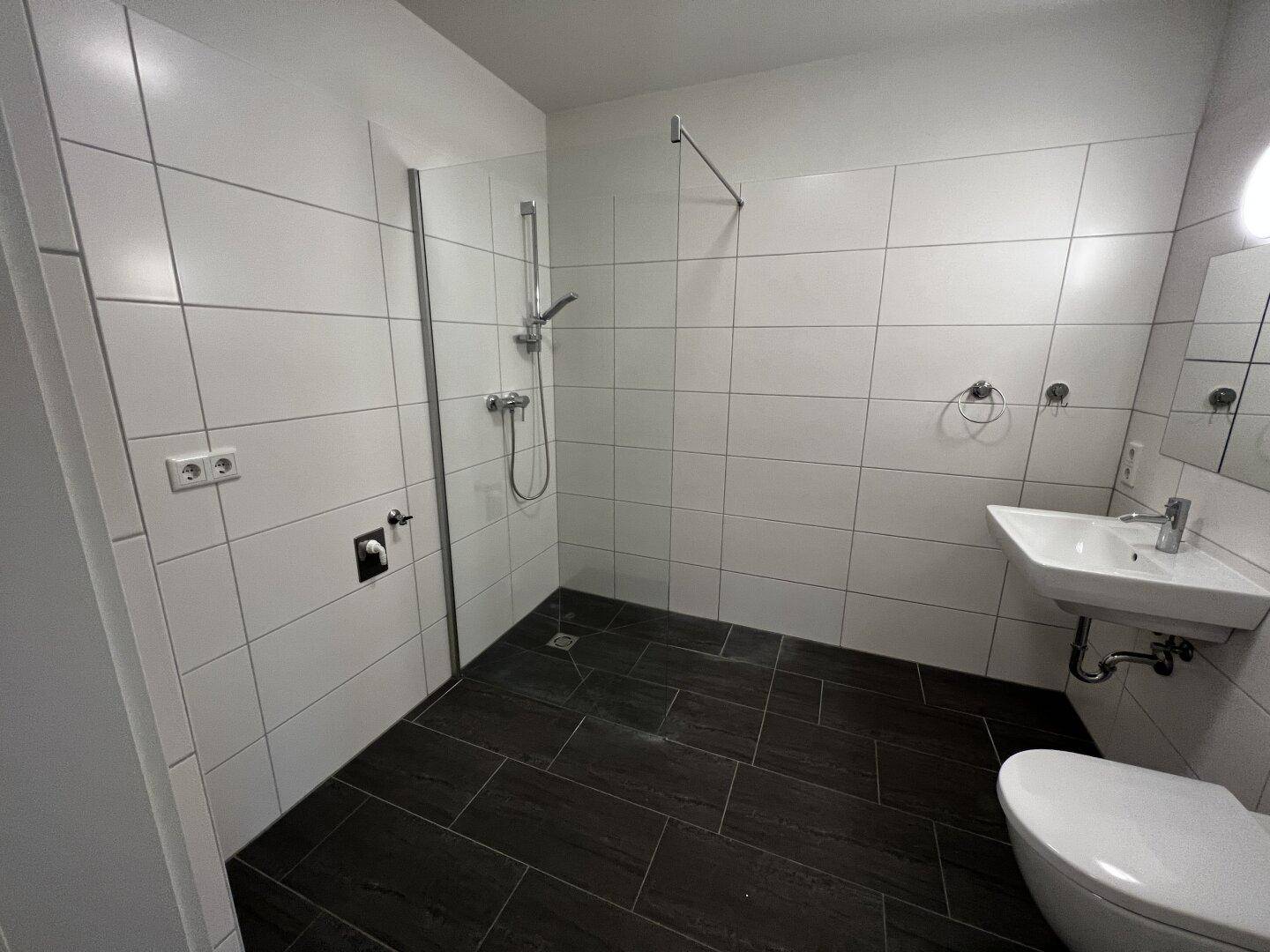 Modernes Badezimmer mit weißen Fliesen, dunklem Boden, ebenerdiger Dusche und WC.