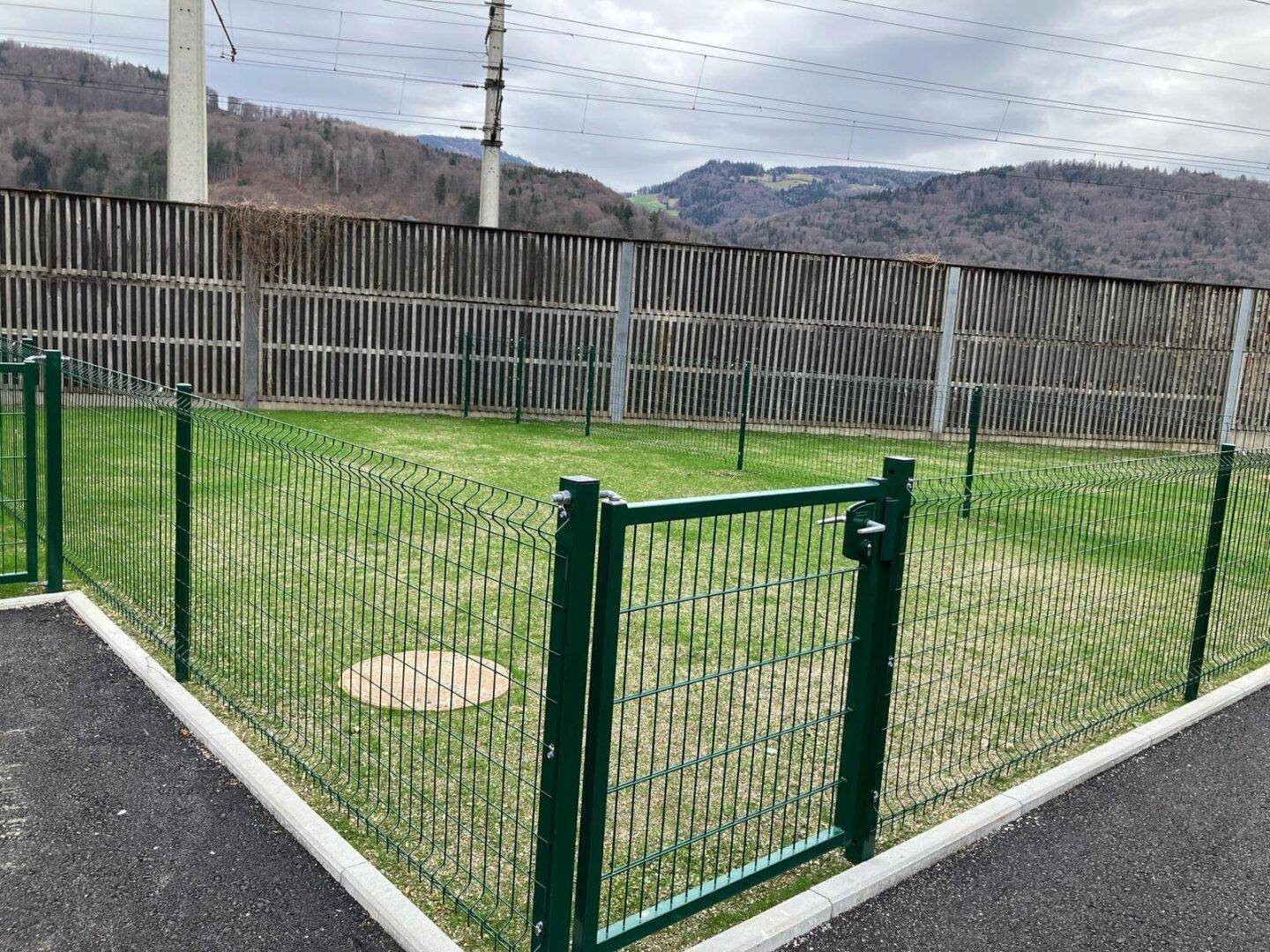 Gepflegte, eingezäunte Rasenfläche neben einer Lärmschutzwand mit Blick auf bewaldete Hügel.
