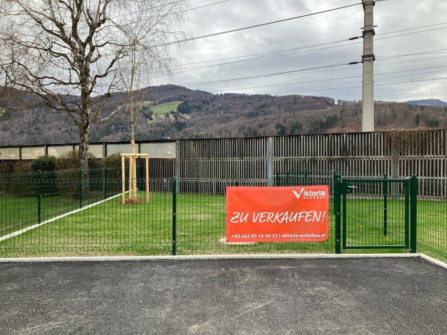 Eingezäunte Grünfläche mit Verkaufsschild, umgeben von einer Lärmschutzwand und bewaldeten Hügeln.