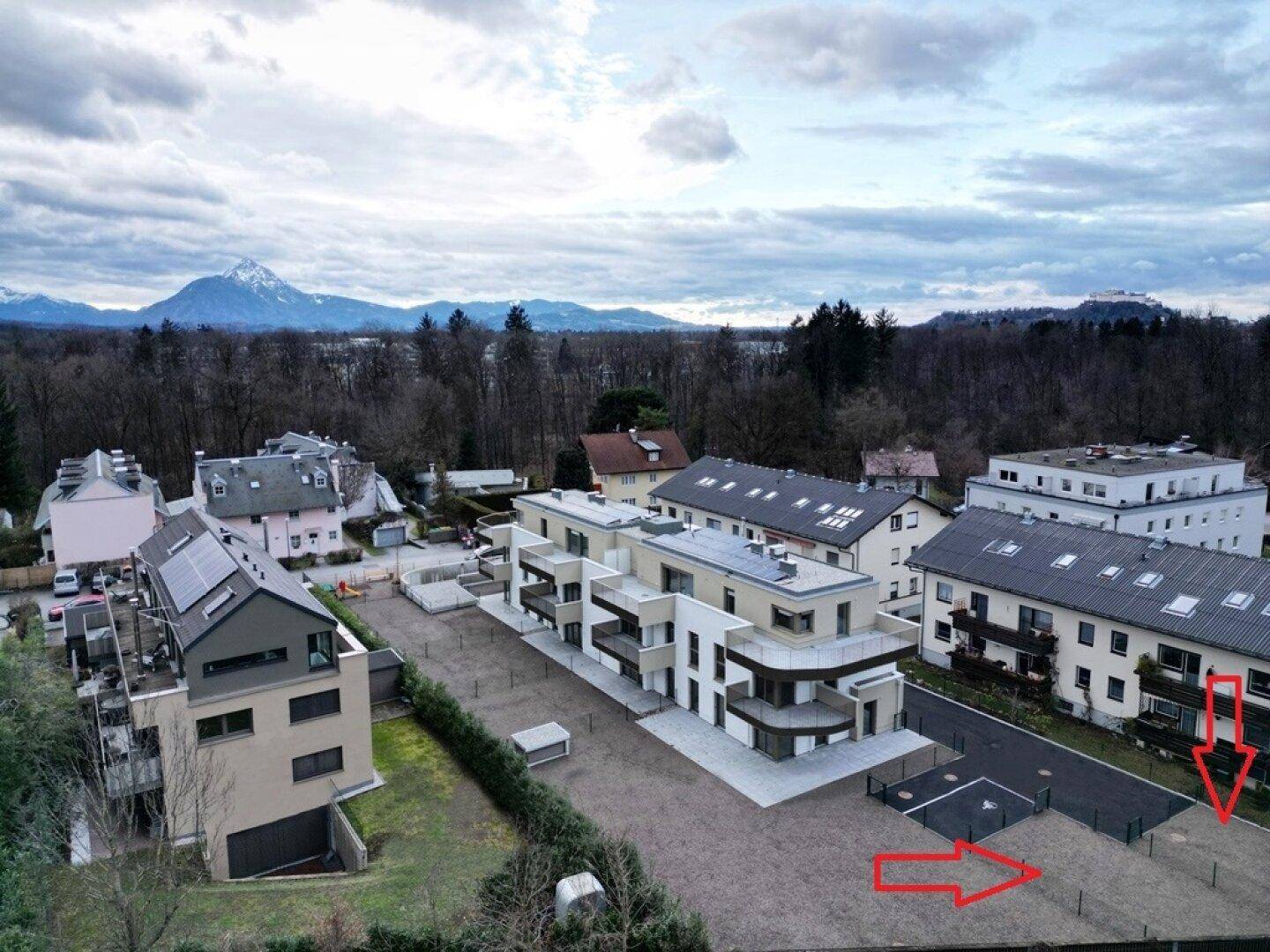 Luftaufnahme einer Wohnsiedlung mit Neubauten und umliegenden Häusern vor einer Bergkulisse.
