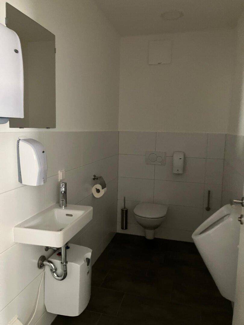 Modernes Badezimmer mit Wand-WC, Urinal und kleinem Waschbecken in Weiß und Dunkelgrau.