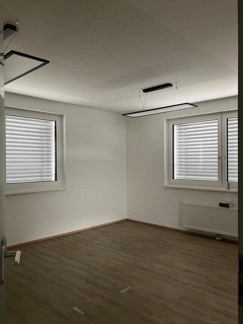 Leerer Büroraum mit zwei Fenstern, Heizkörper und modernen Deckenleuchten.