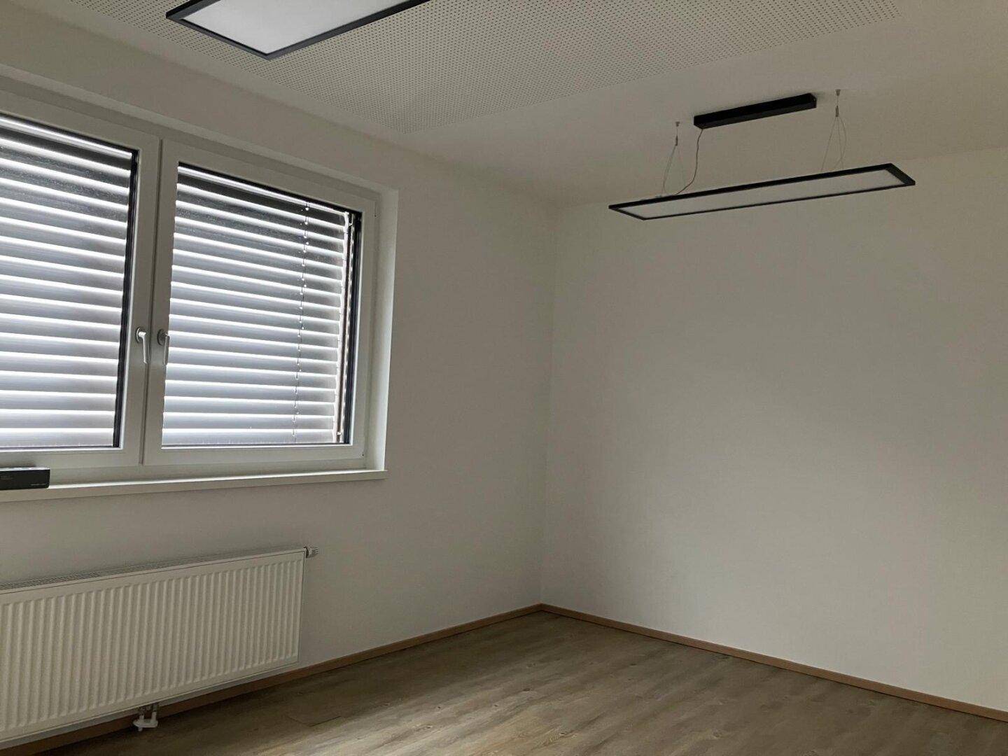 Heller Büroraum mit großem Fenster, Heizkörper und moderner Deckenbeleuchtung.