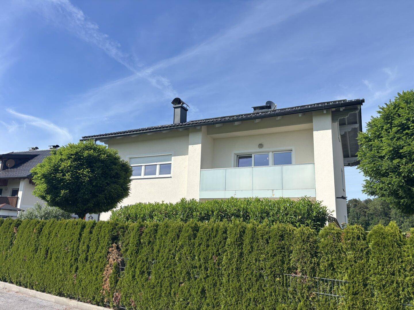 Ansicht des modernen Hauses mit Balkon und einer hohen, grünen Hecke als Grundstücksbegrenzung.