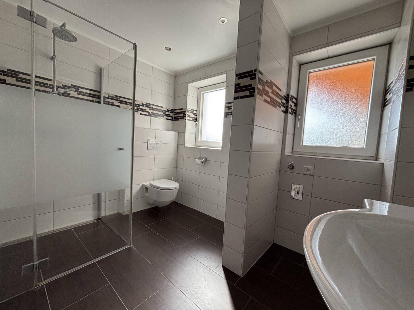 Modernes Badezimmer mit ebenerdiger Dusche, Wand-WC und Fenster, ausgestattet mit dunklen Bodenfliesen.
