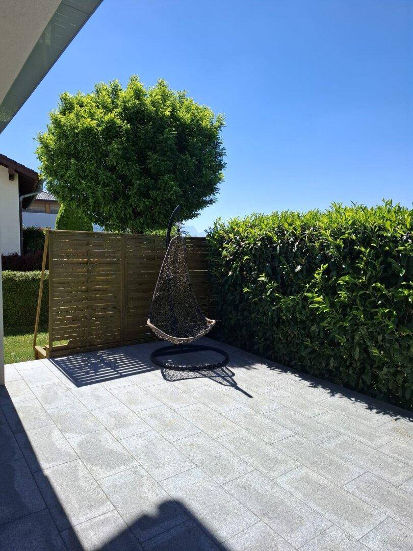 Sonniger Terrassenbereich mit modernen Pflastersteinen, Hängesessel und grüner Hecke.