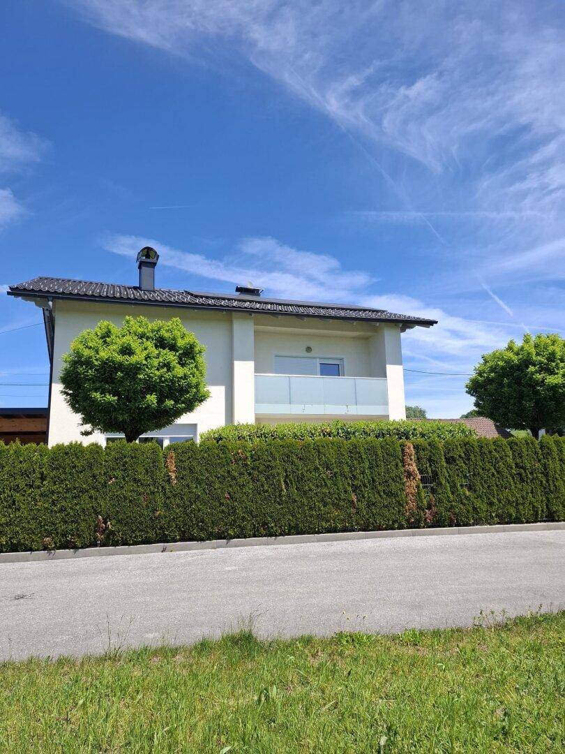 Ansicht des modernen Hauses mit Balkon und einer hohen, grünen Hecke als Grundstücksbegrenzung.