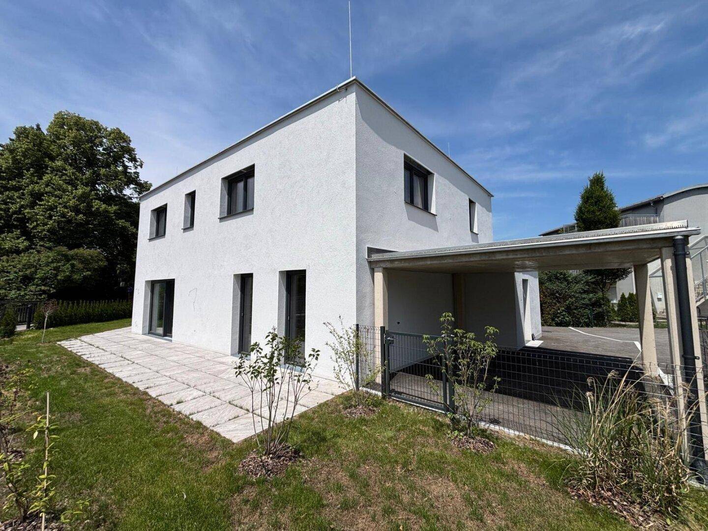 Seitenansicht des modernen Hauses mit gepflasterter Terrasse, Carport und gepflegtem Rasenbereich.