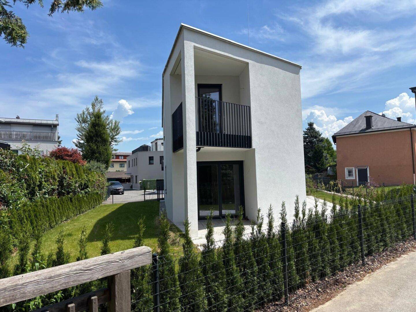 Ansicht des modernen Gebäudes mit Balkon und Eingangsbereich, umgeben von einer grünen Hecke und viel Tageslicht.