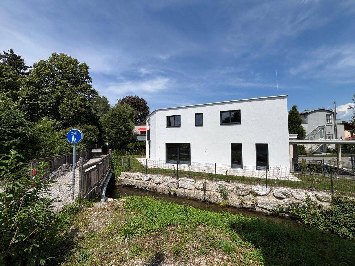 Rückseite des modernen Hauses am Bachlauf mit angrenzender Fußgängerbrücke und grüner Umgebung.