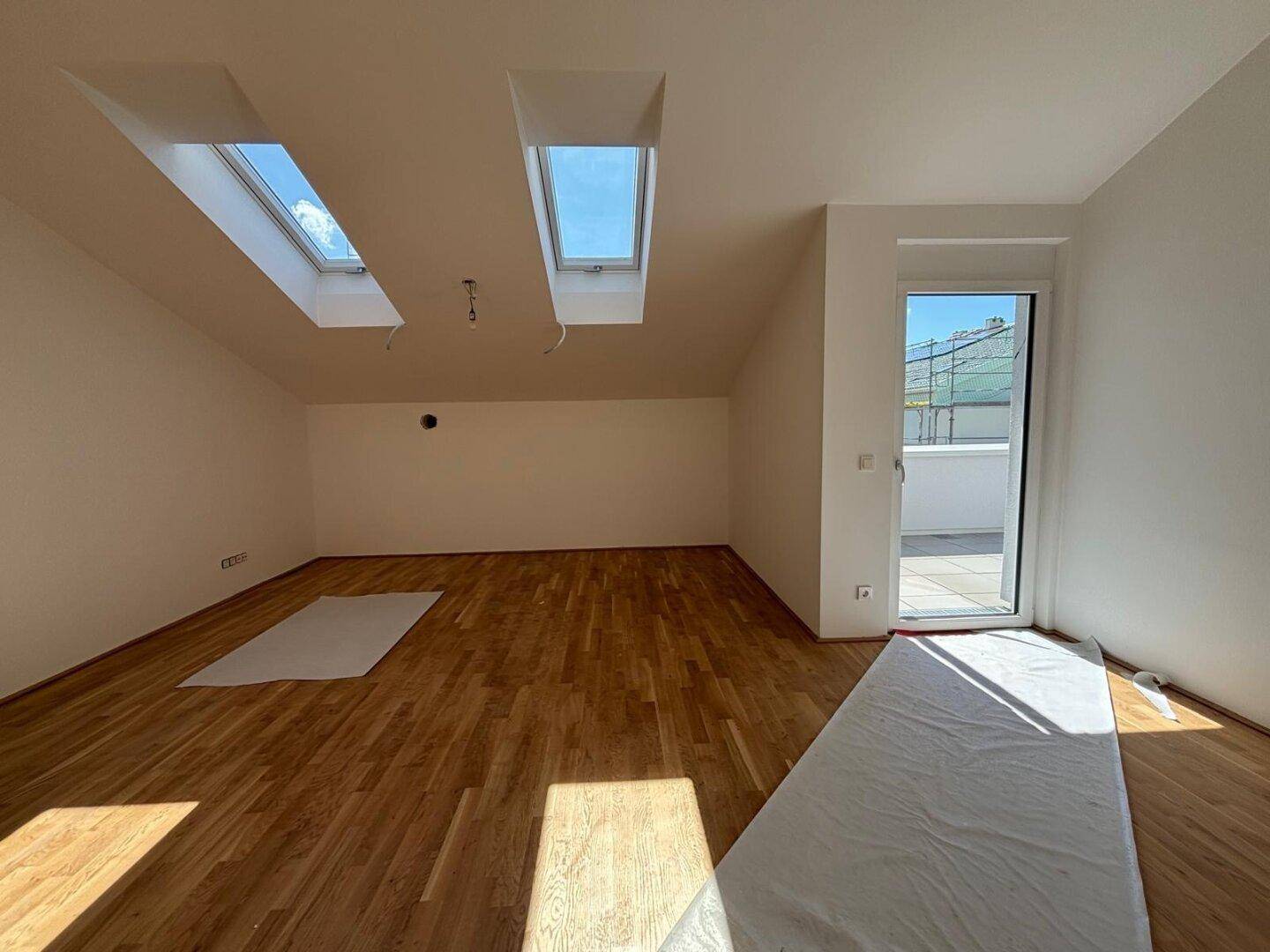 Heller Raum im Dachgeschoss mit Holzboden, Dachfenstern und Zugang zu einem Außenbereich.