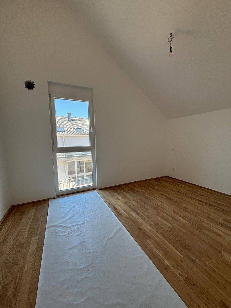 Geräumiger Raum im Dachgeschoss mit Holzboden, schrägen Wänden und einem hohen Fenster.