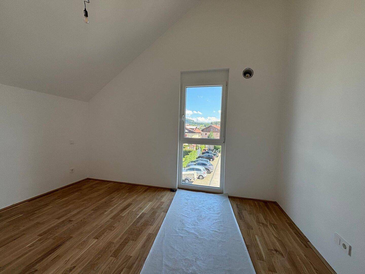 Leeres Zimmer mit Holzboden, schrägen Wänden und einem hohen Fenster mit Blick auf die Umgebung.