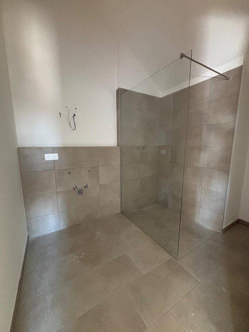 Unfertiges Badezimmer mit grauen Fliesen und einer Glasabtrennung für eine begehbare Dusche.