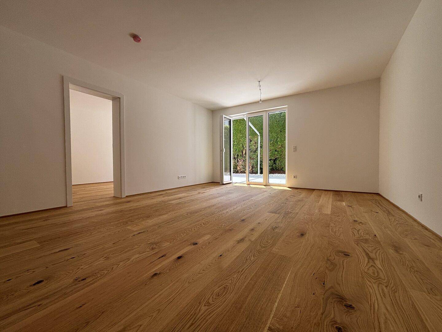 Heller, leerer Raum mit Holzboden und großen Glastüren, die zu einem grünen Außenbereich führen.