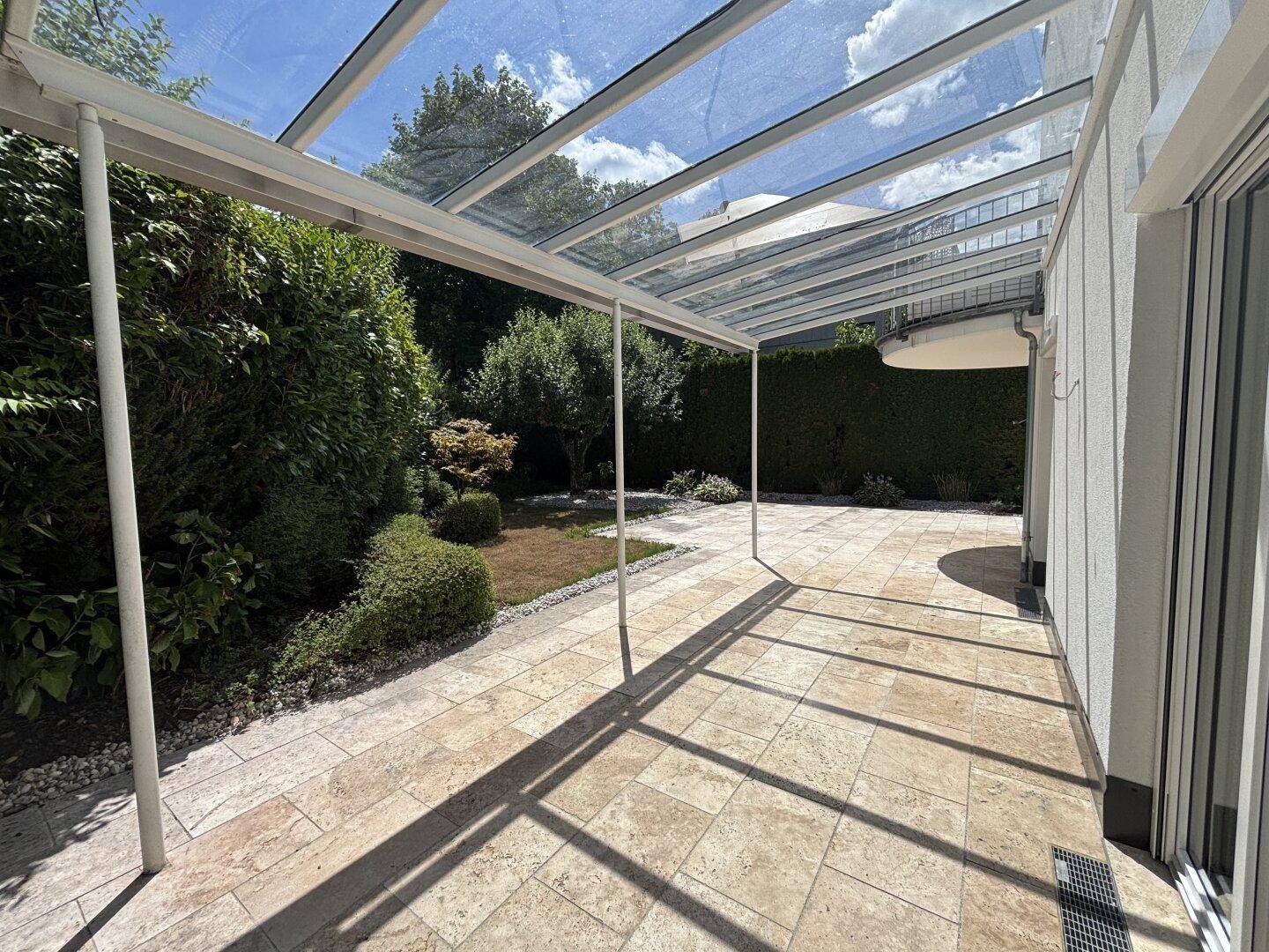 Große, überdachte Terrasse mit Glasdach und hellen Fliesen, umgeben von Grünflächen.