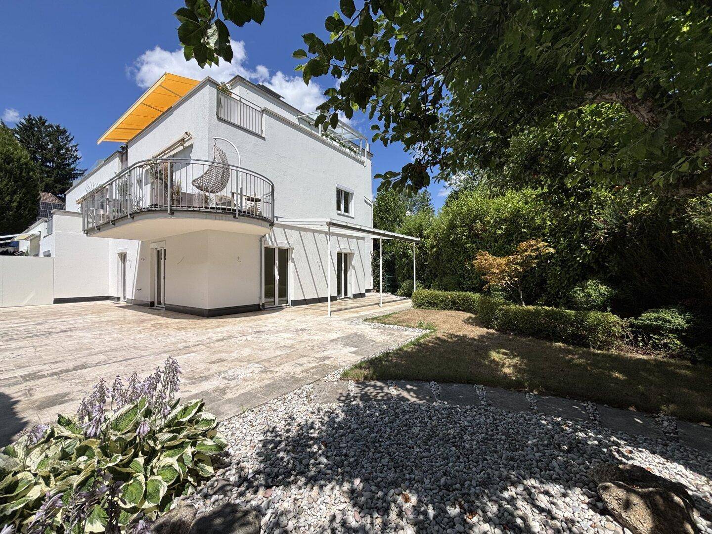 Modernes weißes Gebäude mit geschwungenem Balkon, großer gepflasterter Terrasse und üppigem Garten an einem sonnigen Tag.