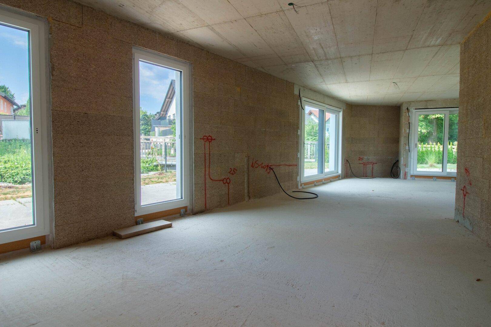 Unfertiger Innenraum im Rohbau mit mehreren Fenster- und Türöffnungen, bereit für den Ausbau.