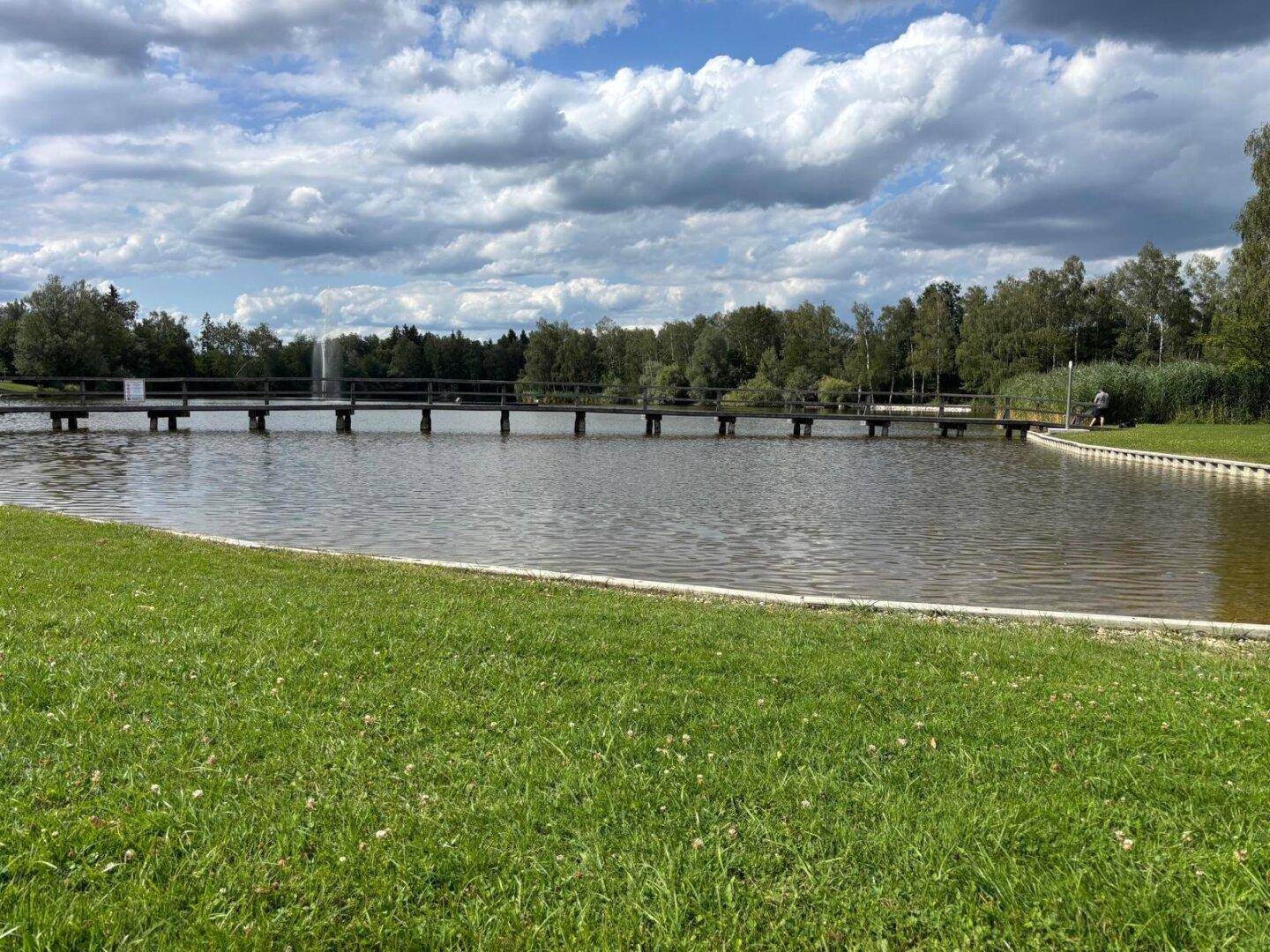 Ein ruhiger See mit einer langen Holzbrücke und einer grünen Liegewiese im Vordergrund.