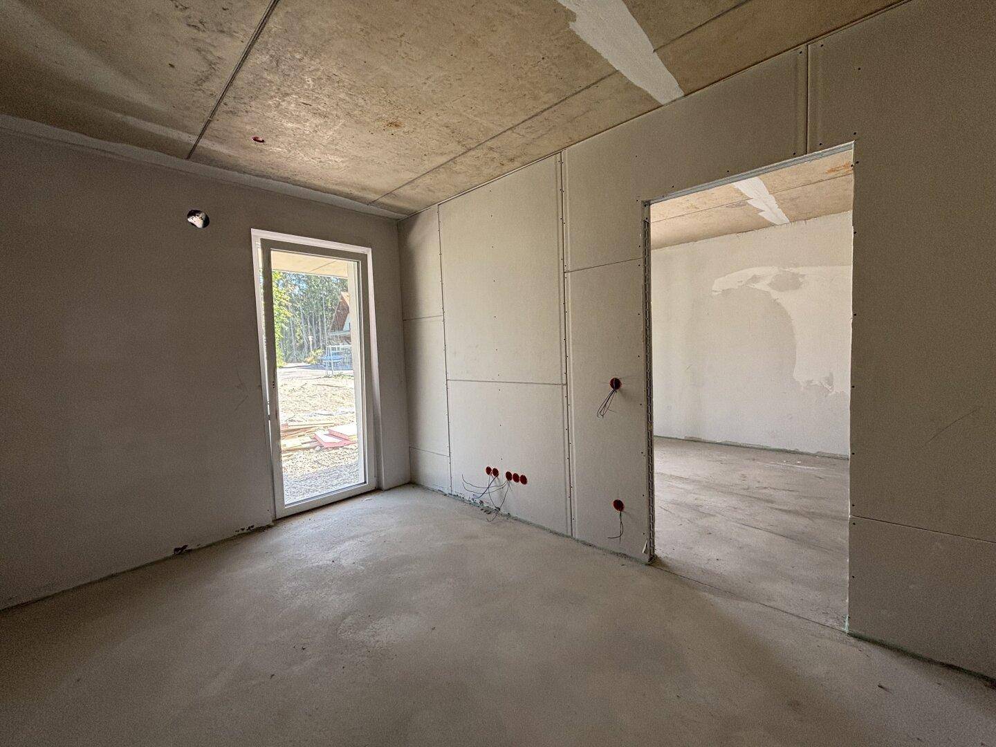 Unfertiger Innenraum im Rohbau mit Betonboden, Gipskartonwänden und Zugang nach draußen durch eine Tür.