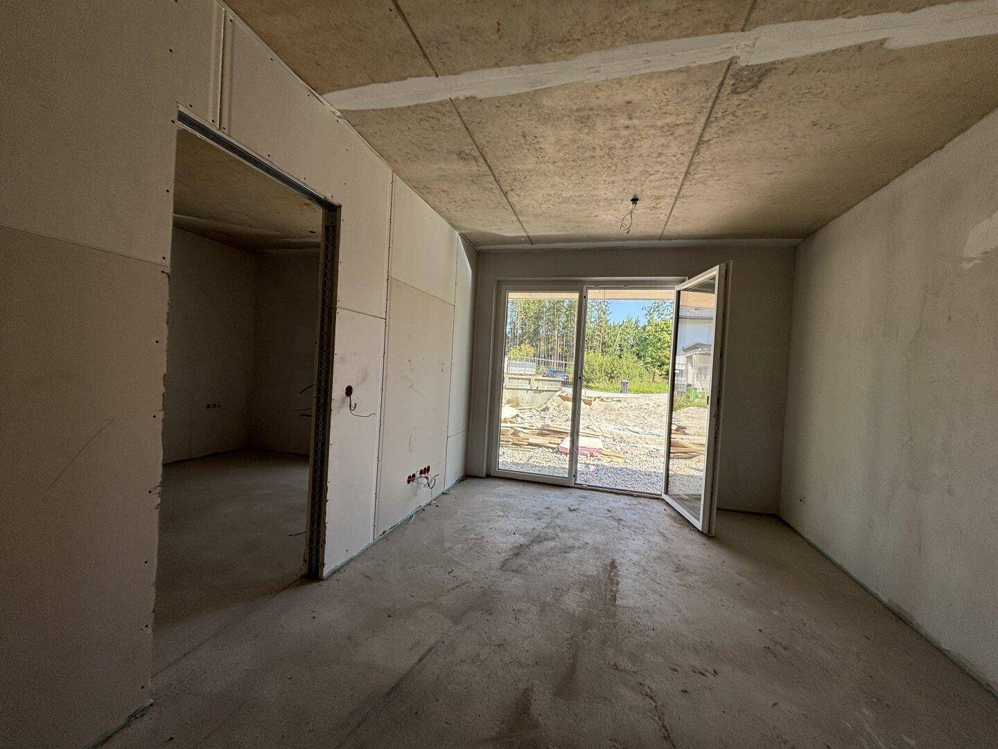 Innenraum im Rohbau mit Betonboden, unverputzten Wänden und einer großen Terrassentür für viel Licht.
