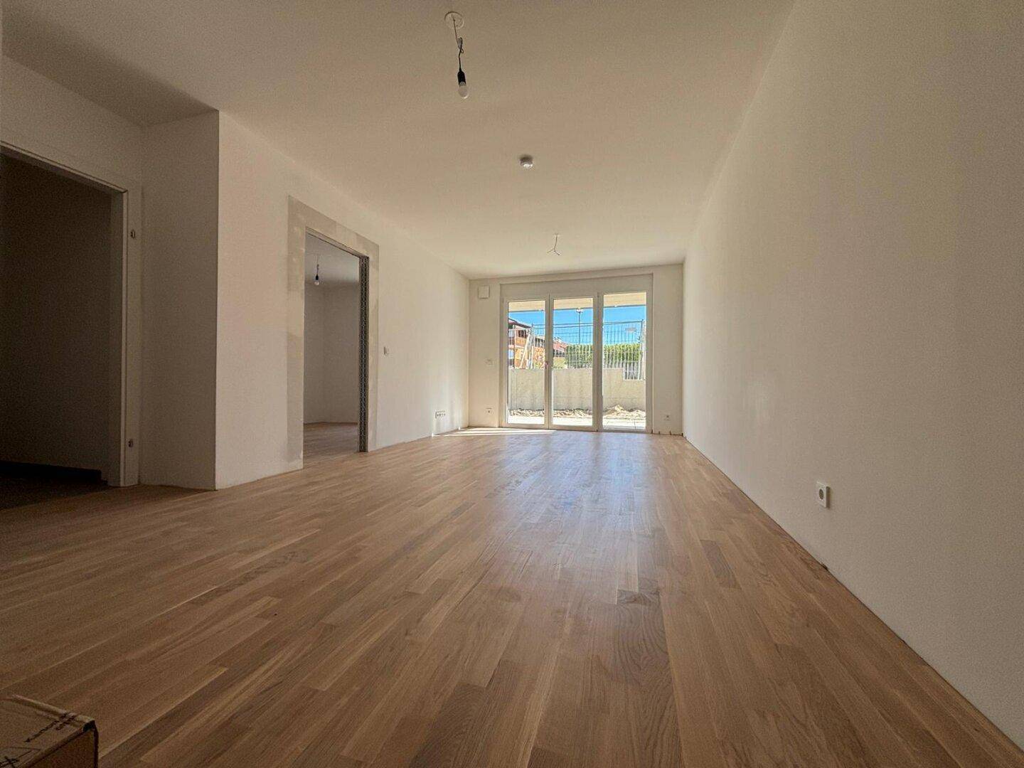 Helles, leeres Wohnzimmer mit neuem Parkettboden und großen Glastüren zum Außenbereich.
