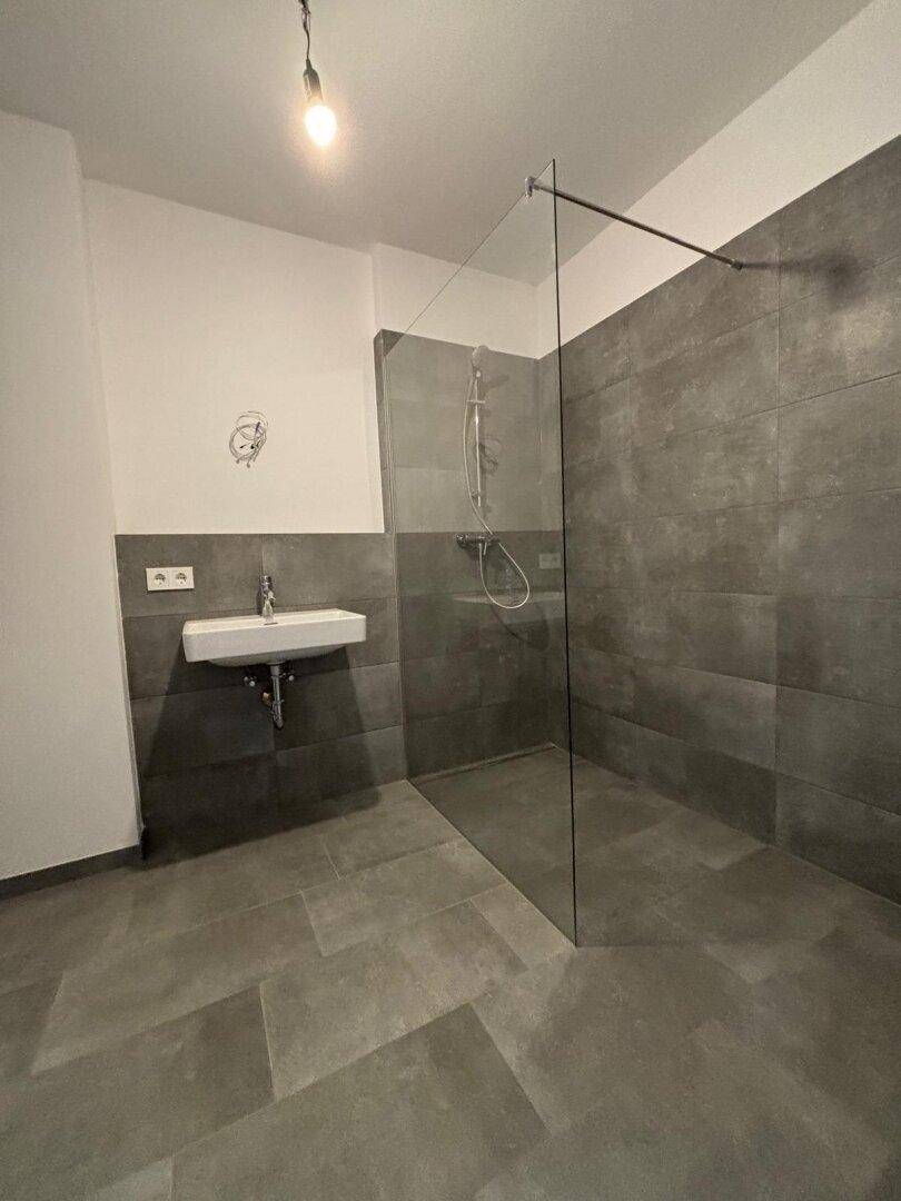 Modernes Badezimmer mit grauen Fliesen, einer begehbaren Dusche und einem Wandwaschbecken.