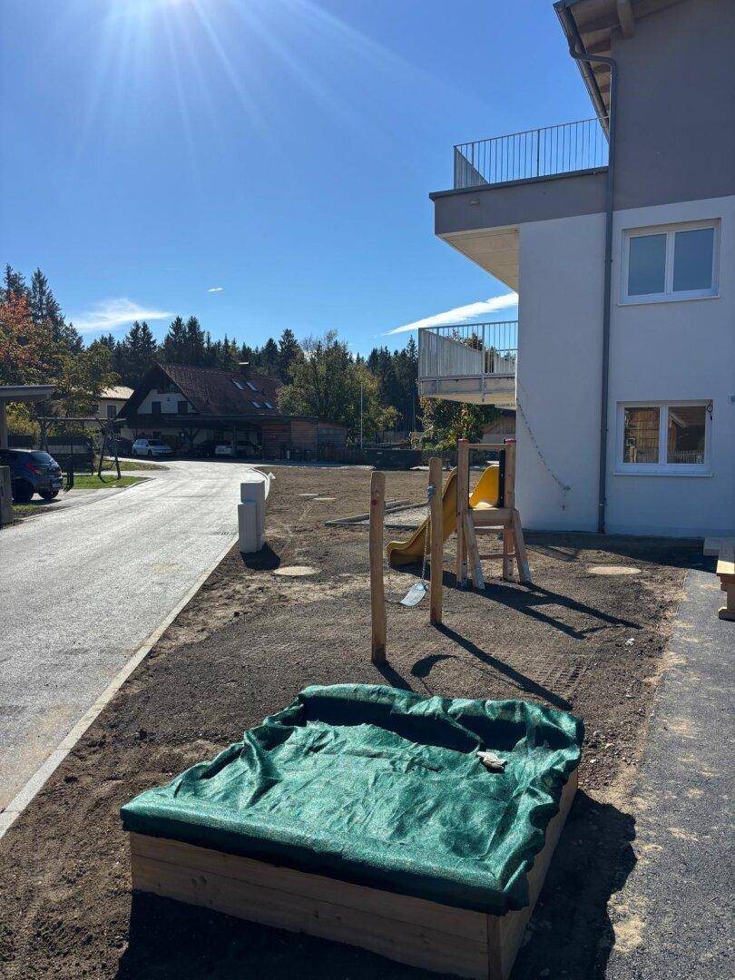 Kinderspielbereich mit Sandkasten und Rutsche neben dem Neubau, umgeben von Bäumen.