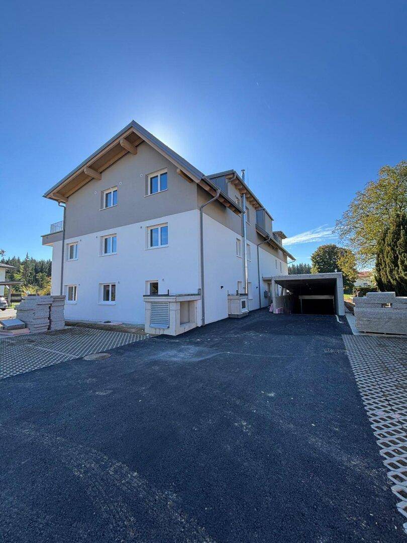 Seitenansicht des Neubaus mit Carport und Zufahrt, umgeben von Bäumen und Grünflächen.
