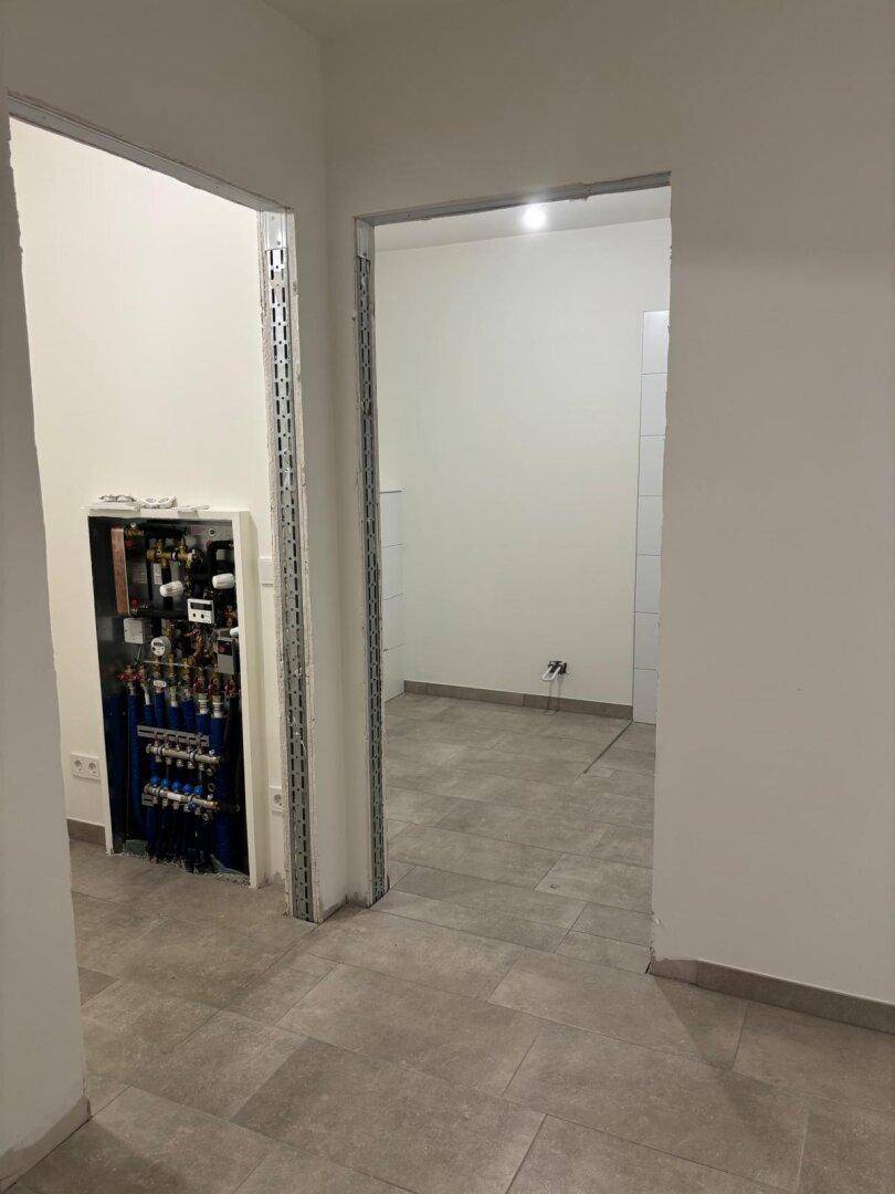 Flur im Rohbau mit Fliesenboden und sichtbarem Verteilerkasten, führt zu weiteren Räumen.