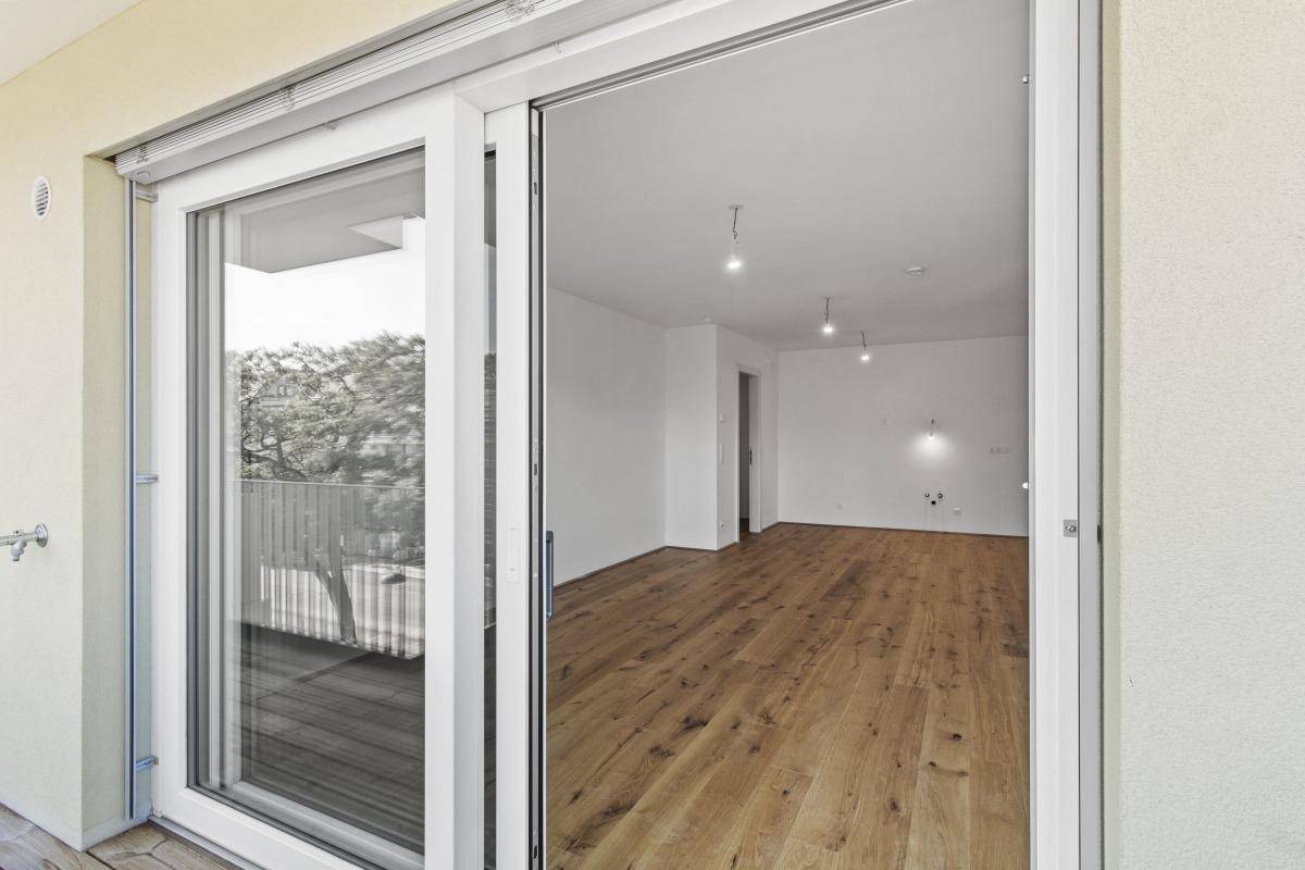 Blick vom Balkon in den hellen, leeren Innenraum mit Holzboden und großen Fenstern.