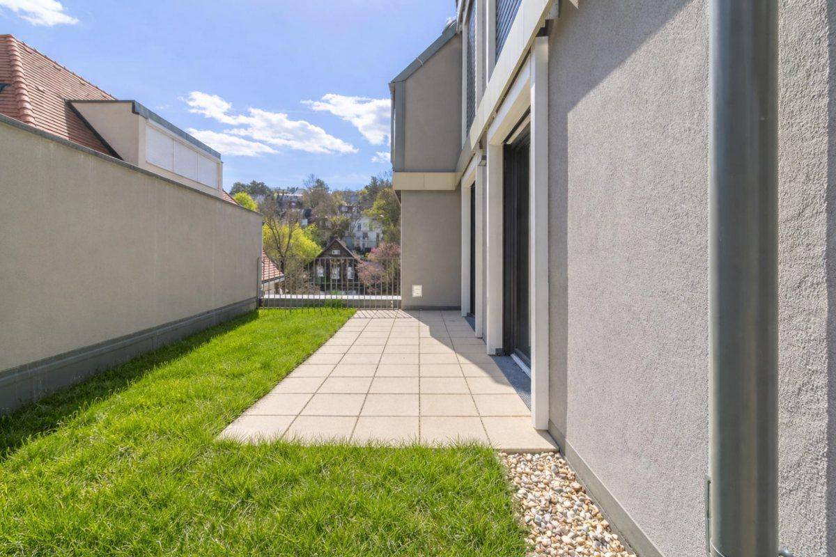 Schmale gepflasterte Terrasse mit Rasenfläche entlang der Gebäudeseite, ideal für Entspannung.