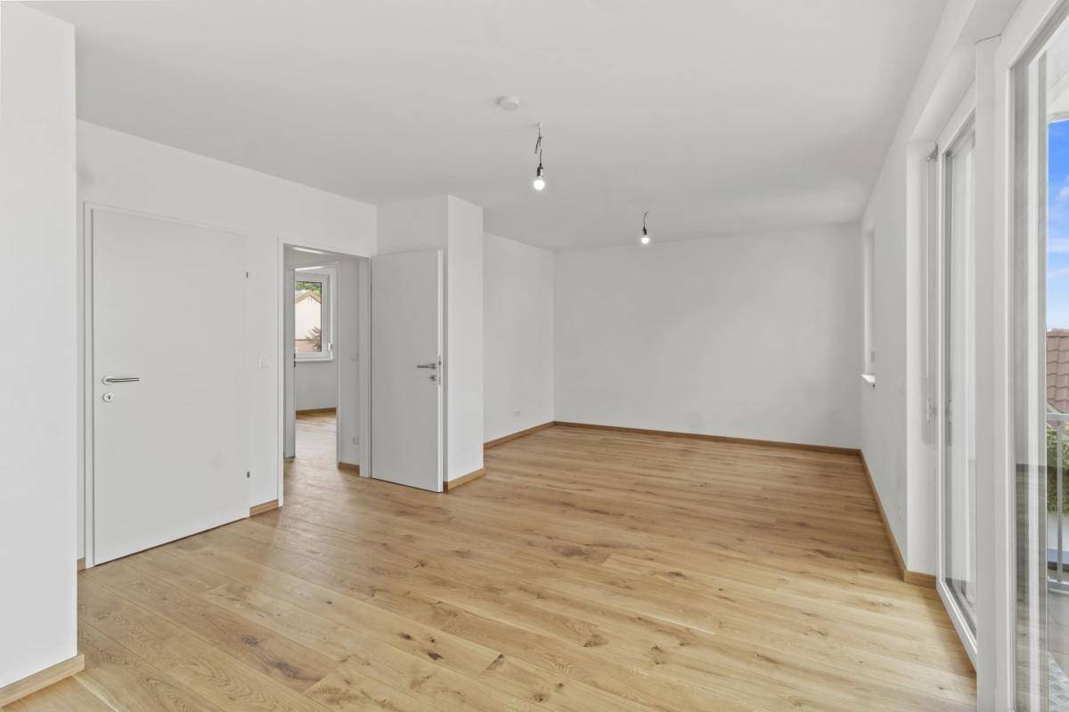 Großzügiges Wohnzimmer mit hellem Parkettboden, weißen Wänden und Balkonzugang.