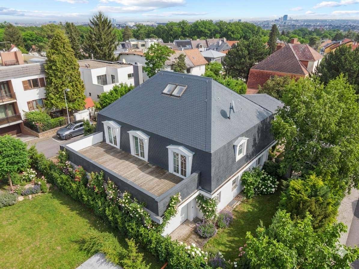 Vogelperspektive eines eleganten Hauses mit dunklem Dach und weitläufiger Terrasse, umgeben von Bäumen.