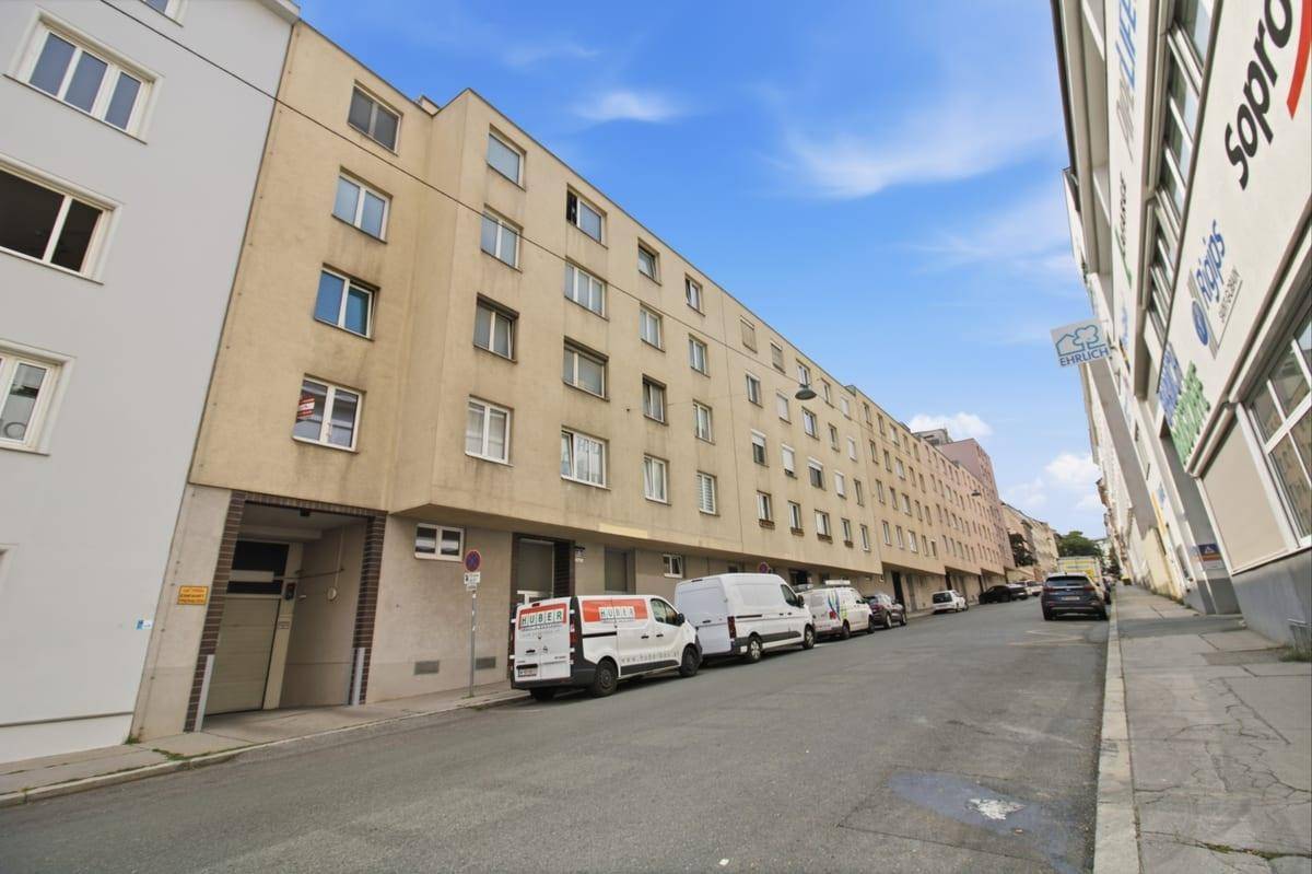 Straßenansicht eines Mehrfamilienhauses mit parkenden Fahrzeugen und Geschäften im Erdgeschoss.
