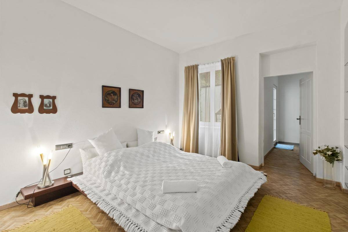 Helles Schlafzimmer mit großem Bett, weißer Bettwäsche, Parkettboden und einem Fenster mit Vorhängen.