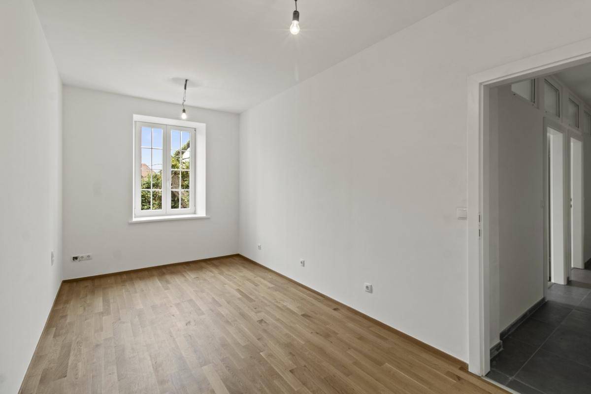 Helles, leeres Zimmer mit Holzdielenboden, weißen Wänden und einem Fenster mit Blick ins Grüne.