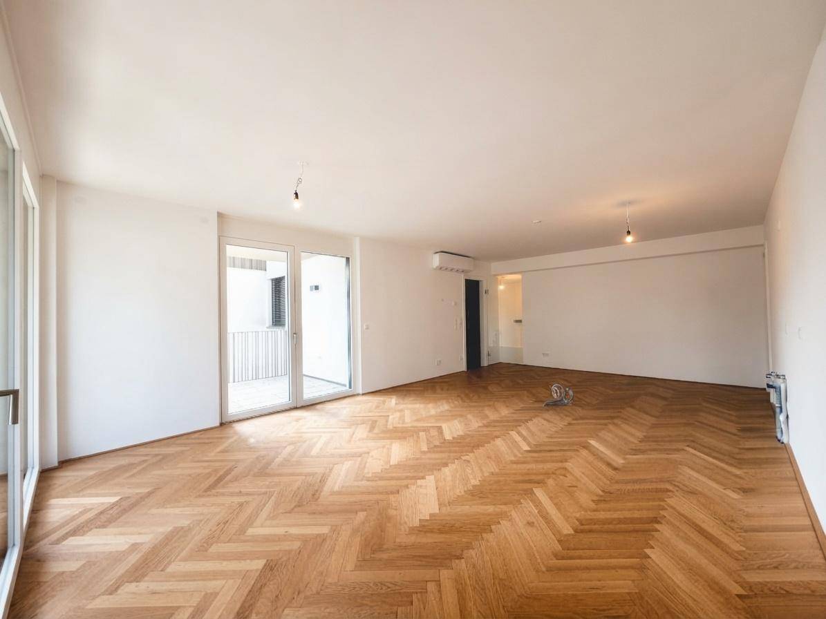 Geräumiges, leeres Wohnzimmer mit Fischgrätparkett, großen Fenstern und Zugang zu einem Balkon.