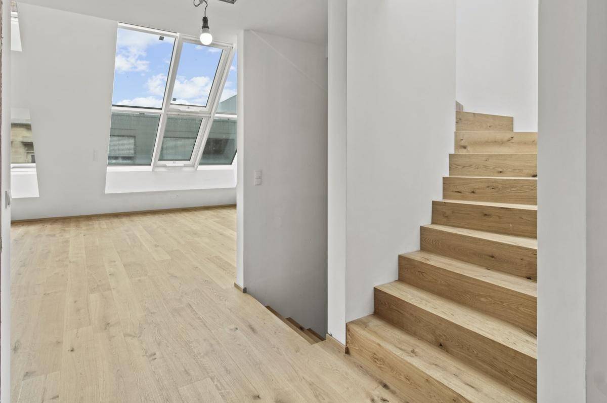 Heller Treppenaufgang mit Holzstufen und großen Dachfenstern, die viel Tageslicht hereinlassen.