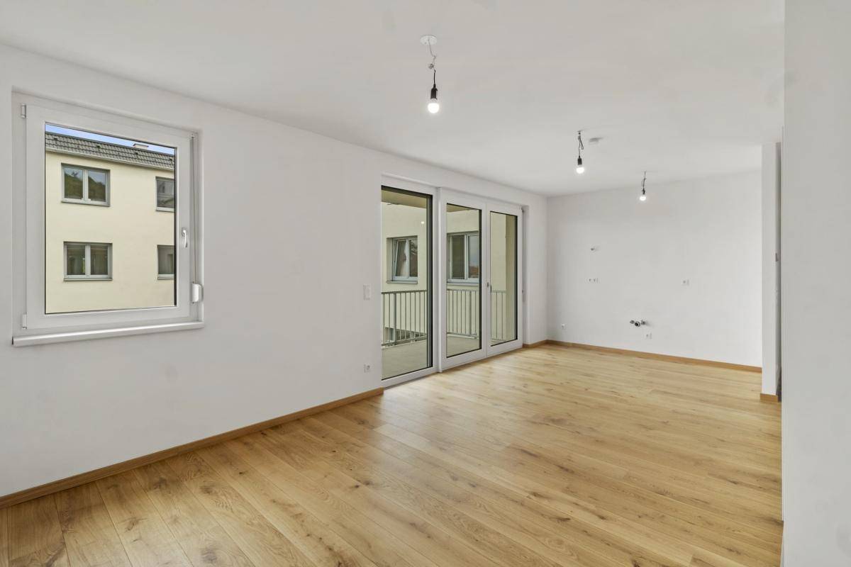 Geräumiges Wohnzimmer mit hellem Parkettboden, großen Fenstern und Balkonzugang.